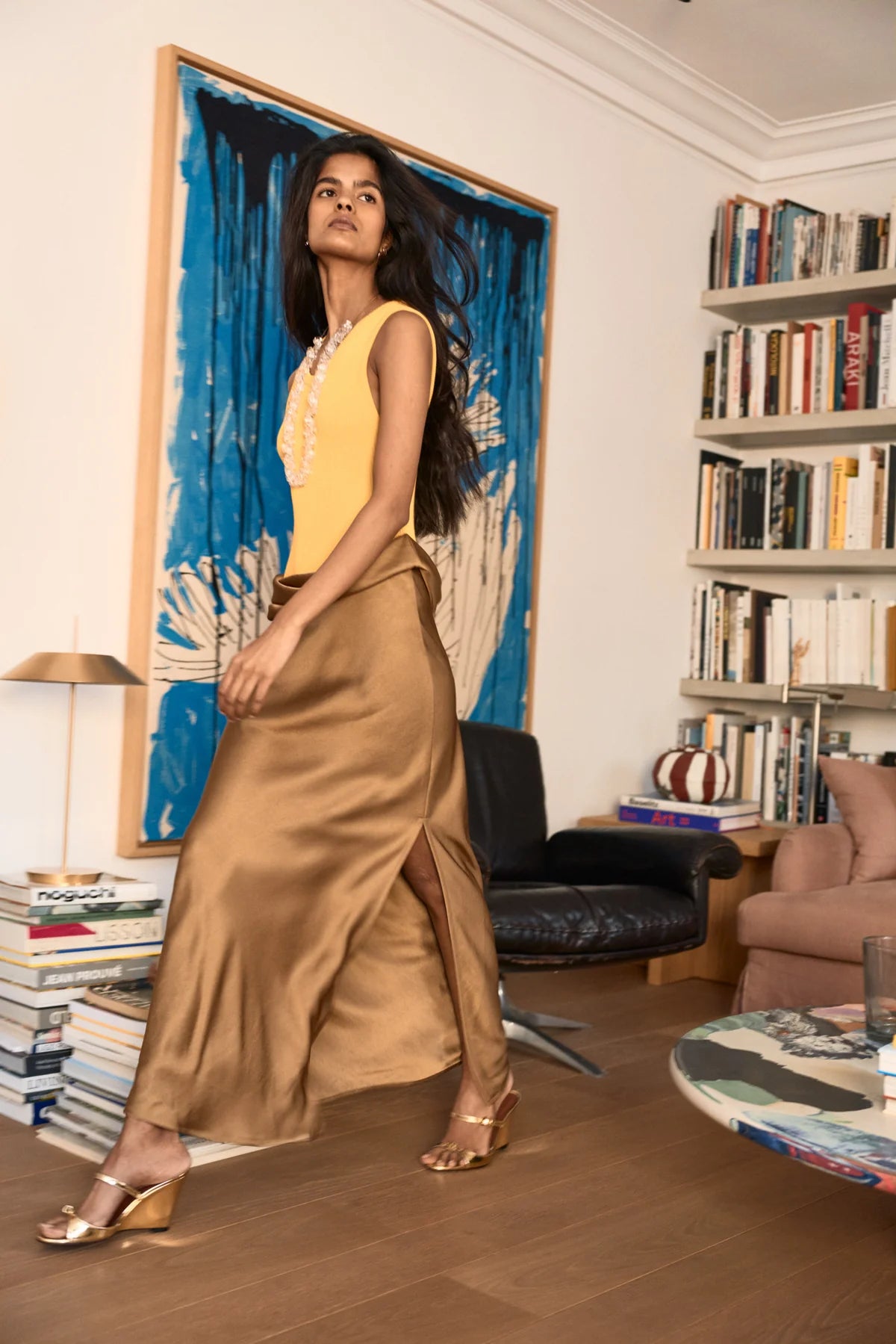 Fashion model in a yellow top and gold skirt standing in a stylish living room.